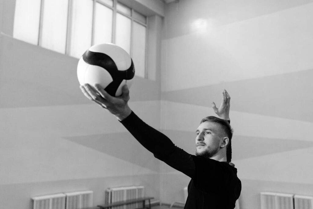 Caucasian man serving a volleyball in an indoor game setting, black and white photo.